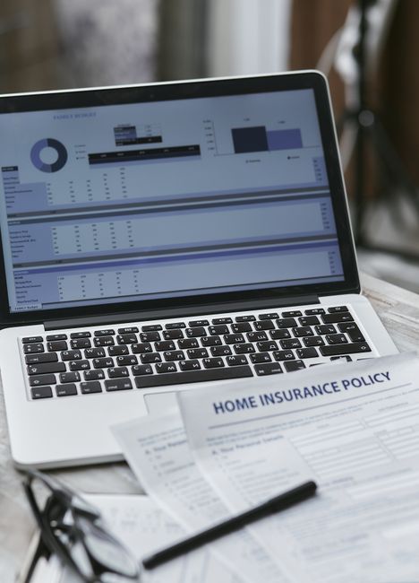 A close-up of a laptop displaying various charts and graphs on its screen, placed on a wooden desk alongside printed documents related to home insurance policies, including one titled 'Home Insurance 