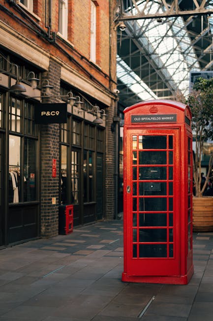 The image depicts a classic red British telephone box situated on a paved walkway outside a building with large glass windows and a brick facade, typical of urban London architecture. The telephone bo