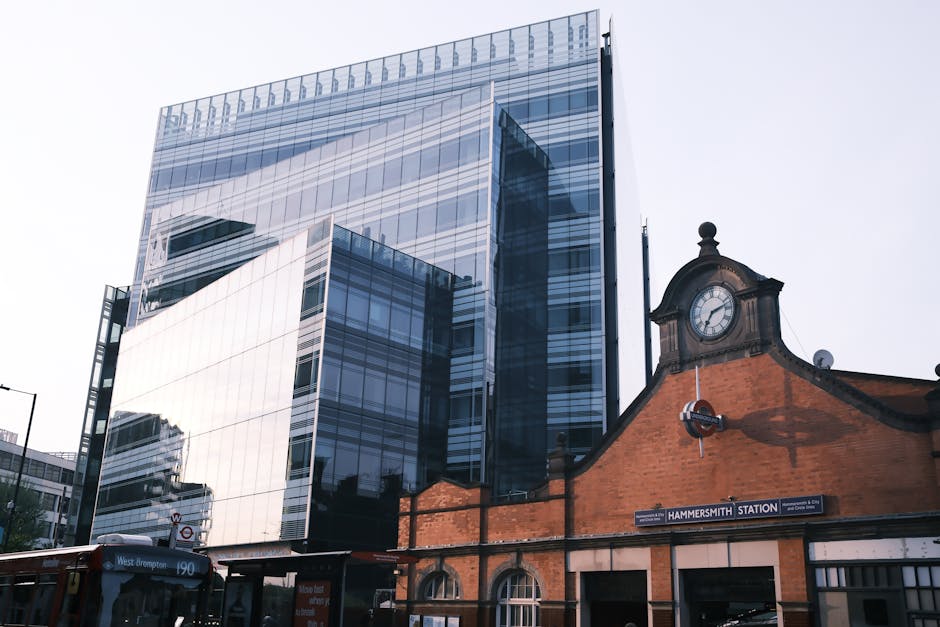 The image depicts a classic red British telephone box situated on a paved walkway outside a building with large glass windows and a brick facade, typical of urban London architecture. The telephone bo